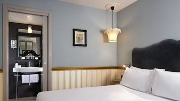 Et hotelværelse med seng, natbord, lampe på væggen, et billede i ramme, og et badeværelse med vask og håndklædestang.