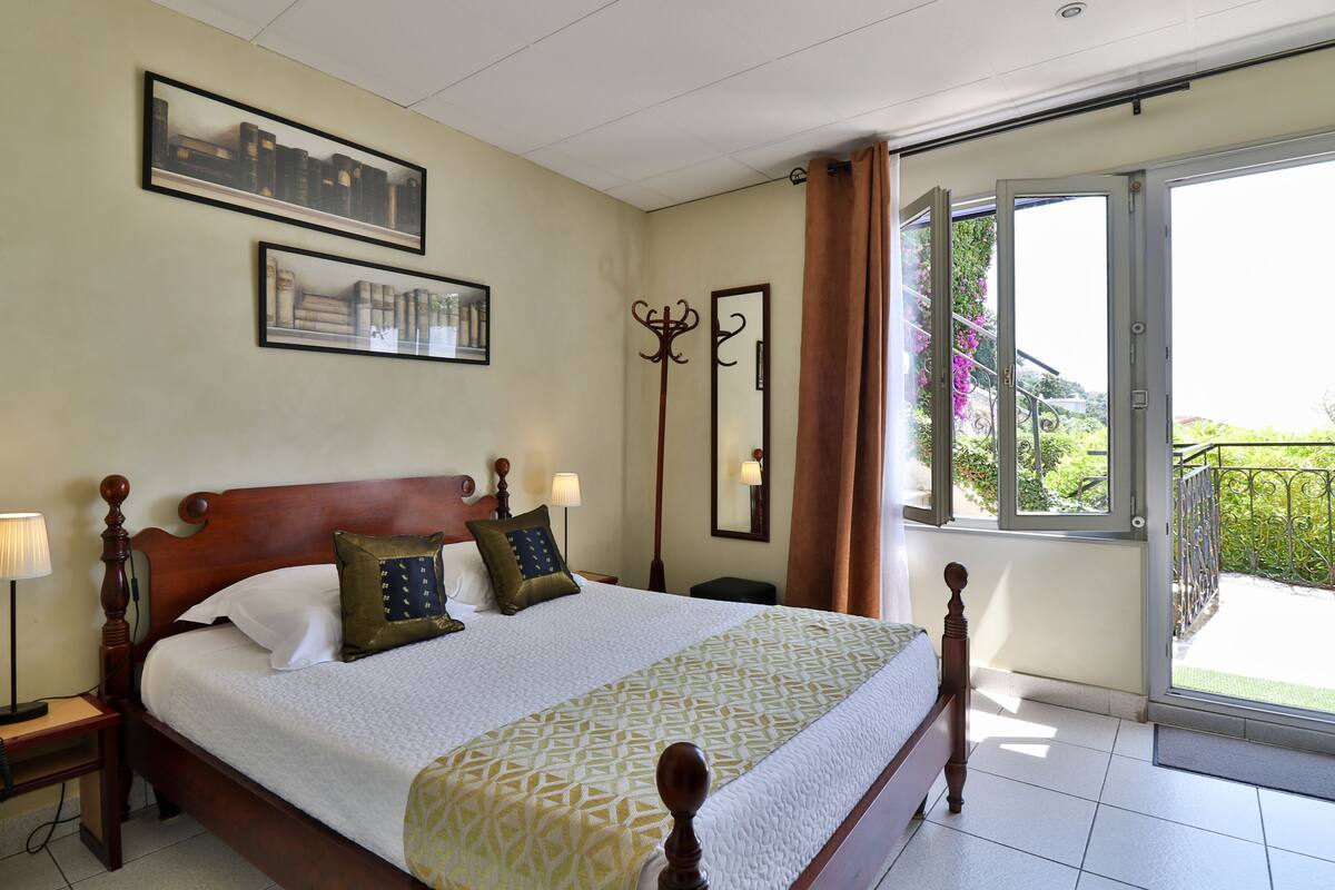 A neatly made bed with a patterned bedspread, a wooden headboard, and two pillows. There are framed pictures on the wall, a nightstand with a lamp, and a large window with curtains.