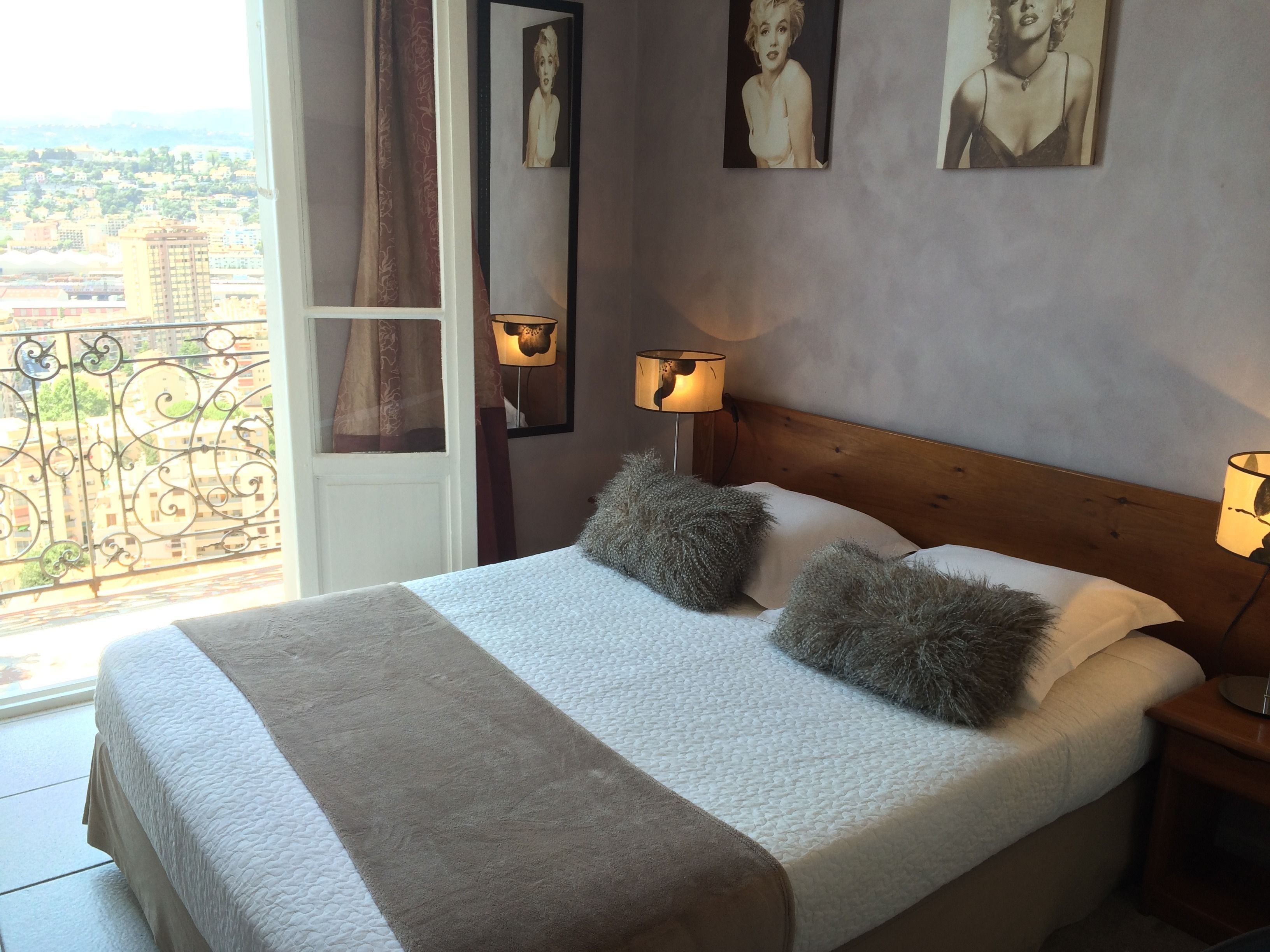 A bedroom with a bed, two pillows, a nightstand, two wall-mounted lamps, and a view of the city through a balcony door.