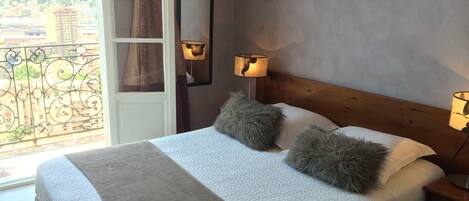 A bedroom with a bed, two pillows, a nightstand, two wall-mounted lamps, and a view of the city through a balcony door.