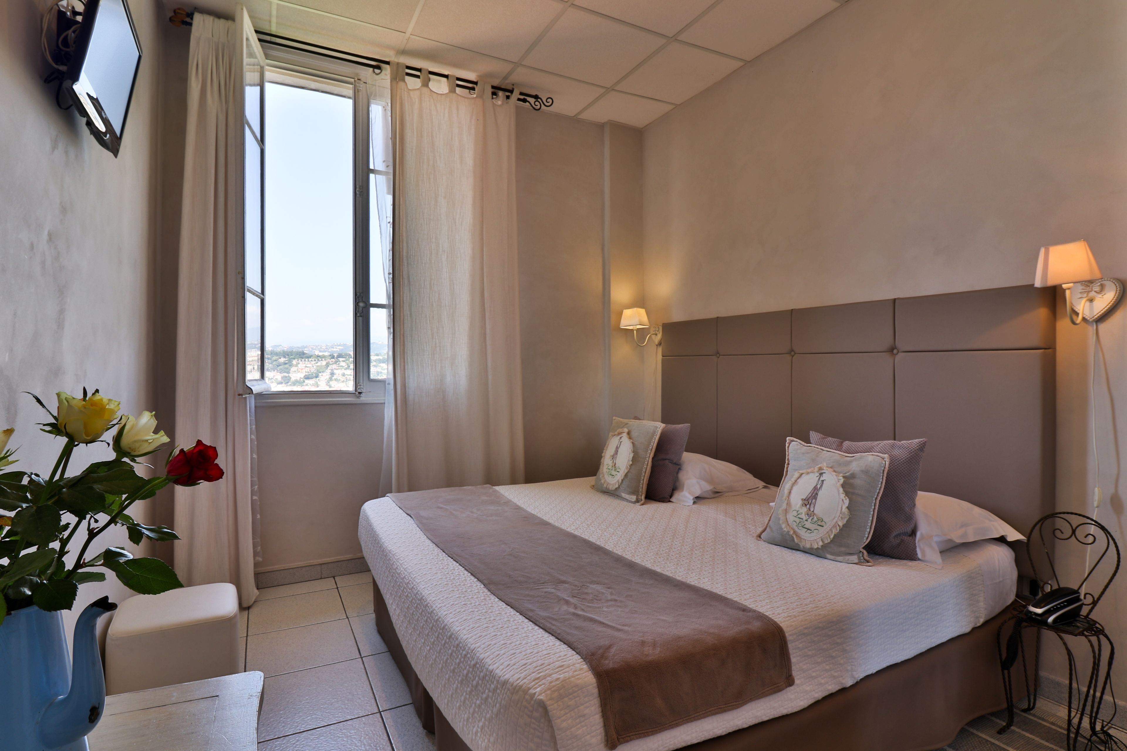 A bedroom with a large bed, a bedside table, a lamp, and a view of the city through a window.