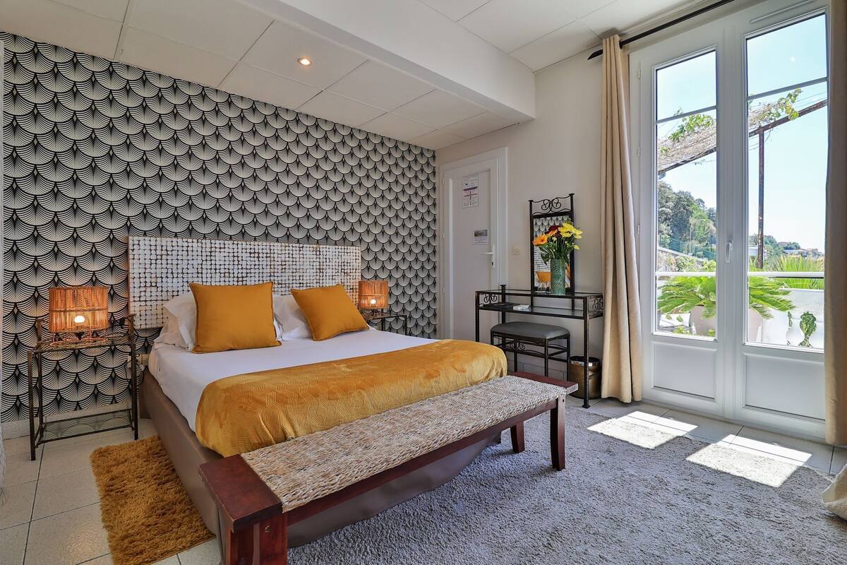 A bedroom with a patterned headboard, a bed with two pillows, a bedside table with a lamp, a bench, and a view of greenery through a window.