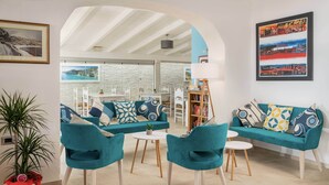 Een moderne woonkamer met turquoise banken, een bijzettafeltje en stoelen. Er is een boekenkast, een plant en een ingelijste foto aan de muur.