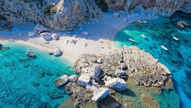 Luchtbeeld van een strand met helder turquoise water, rotsformaties en boten.