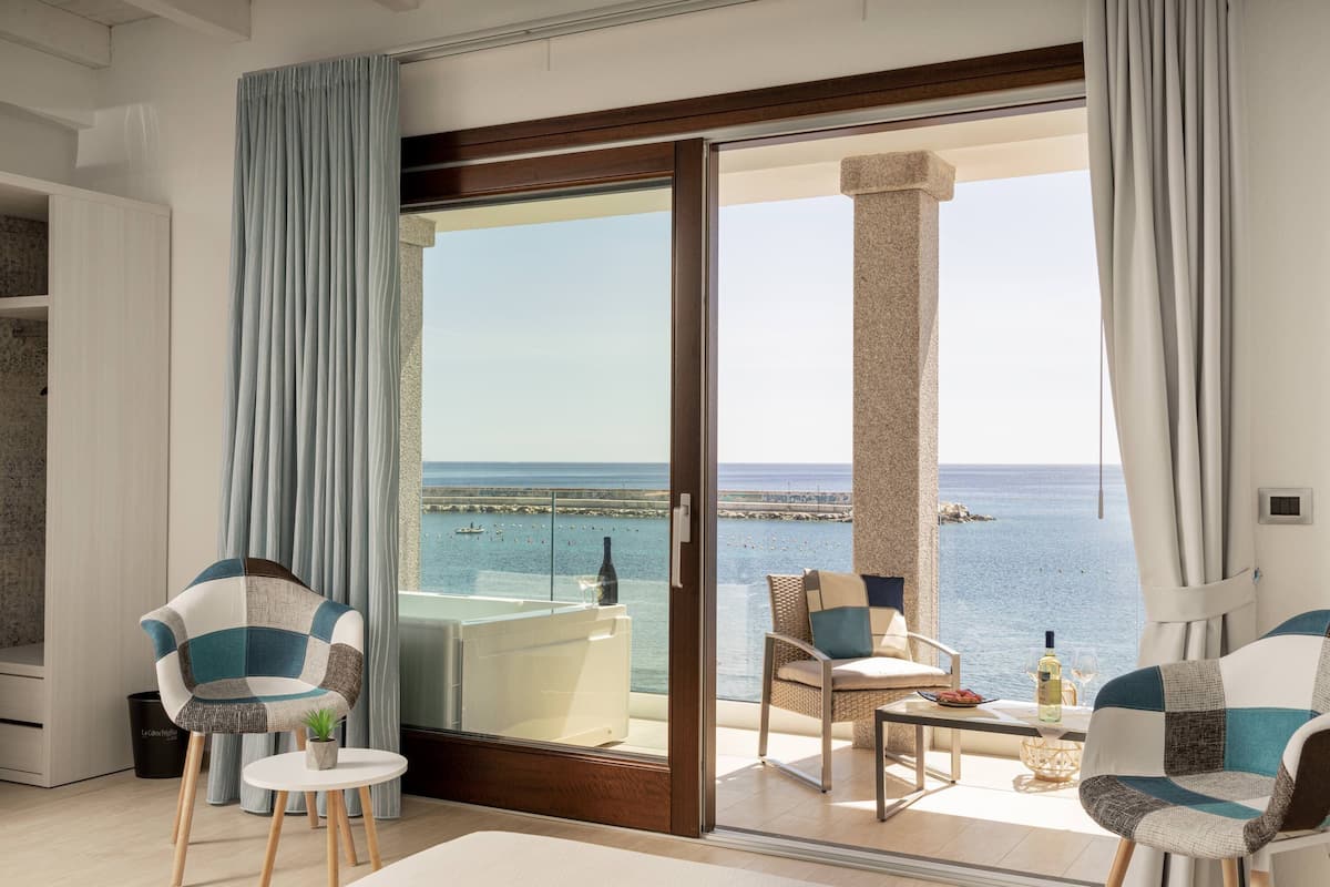 Ein modernes Zimmer mit einer großen Schiebeglasdoor zum Balkon mit Meerblick. Zwei Polsterstühle und ein kleiner runder Tisch mit Flasche und Glas darauf.