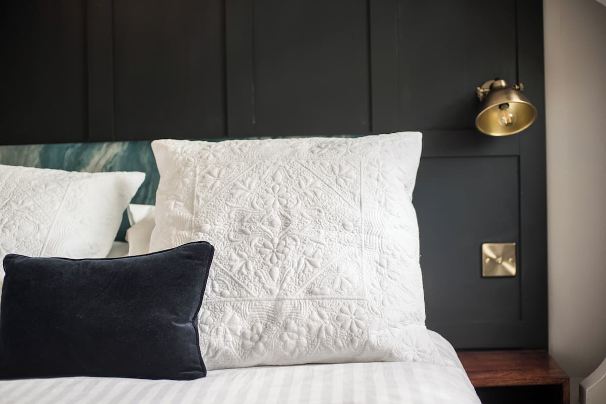 A neatly made bed with white and dark pillows, a dark headboard, and a brass wall sconce.