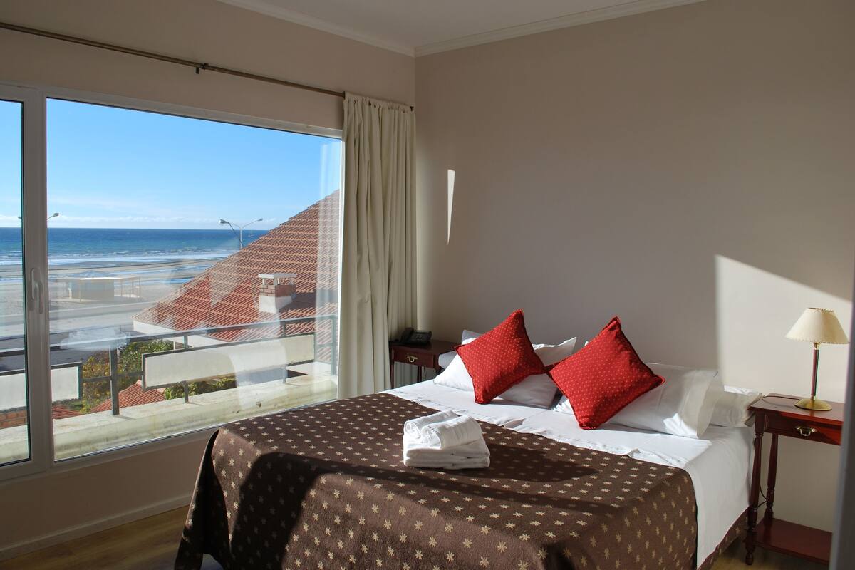 Habitación de hotel con un amplio ventanal que ofrece vistas a la playa y edificios. La habitación tiene una cama con colcha marrón estampada y almohadas rojas.
