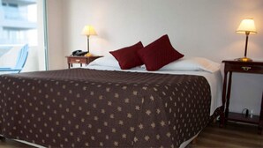 Habitación de hotel con una cama grande, dos mesitas de noche con lámparas, una silla y vistas al exterior.