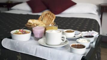 Uma bandeja de café da manhã com croissants, cereais, café e diversas geleias, sobre uma cama com edredom escuro estampado e almofadas vermelhas.