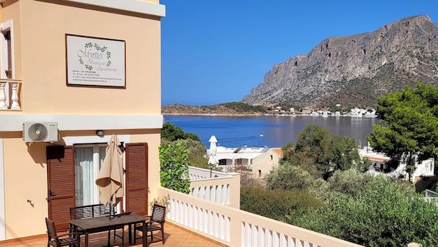 Un immeuble situé sur la côte, doté d’un balcon offrant une vue sur la mer, avec une montagne en arrière-plan, et une enseigne indiquant « Myrtis Apartments ».