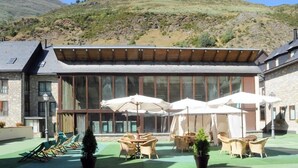 A building with a large glass facade, outdoor seating area with chairs and umbrellas, and a green lawn. - Hotel y Apartamentos Vilagaros (Naut Aran)