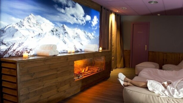 A room with a large TV displaying a mountain scene, a wooden fireplace, and a person relaxing on a sofa. - Hotel y Apartamentos Vilagaros (Naut Aran)