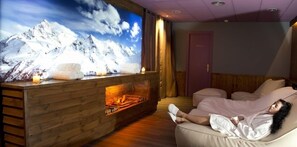 Ein Zimmer mit einem großen Fernseher, der eine Berglandschaft zeigt, einem Kamin aus Holz und einer Person, die entspannt auf einem Sofa sitzt.