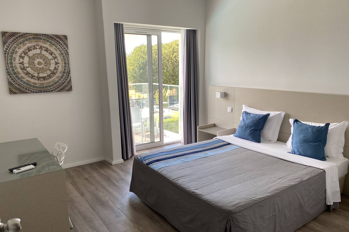 A modern hotel room with a large bed, a desk, and a view of the outdoors through a sliding glass door.