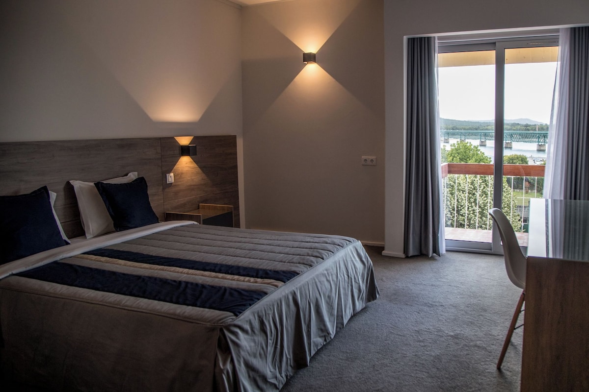 A hotel room with a bed, a desk, a chair, and a view of a bridge and greenery through a large window.