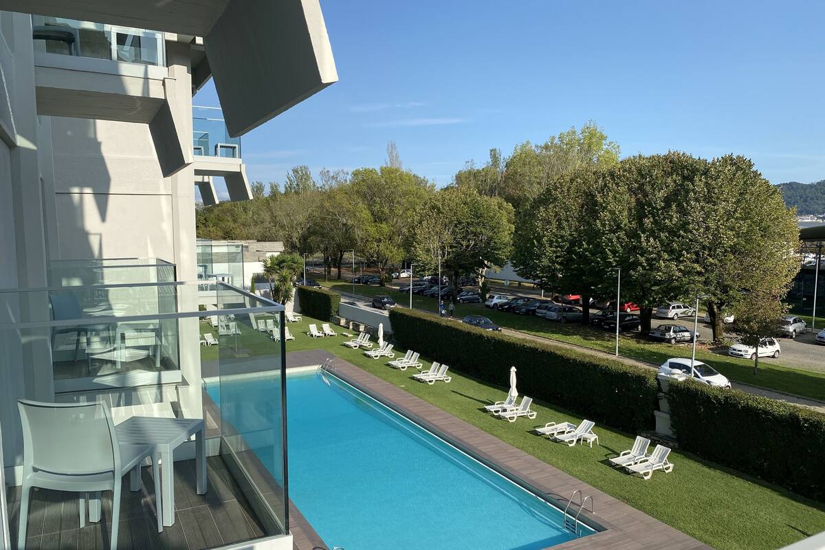 A modern building with a swimming pool, lounge chairs, and a view of a parking area with trees.