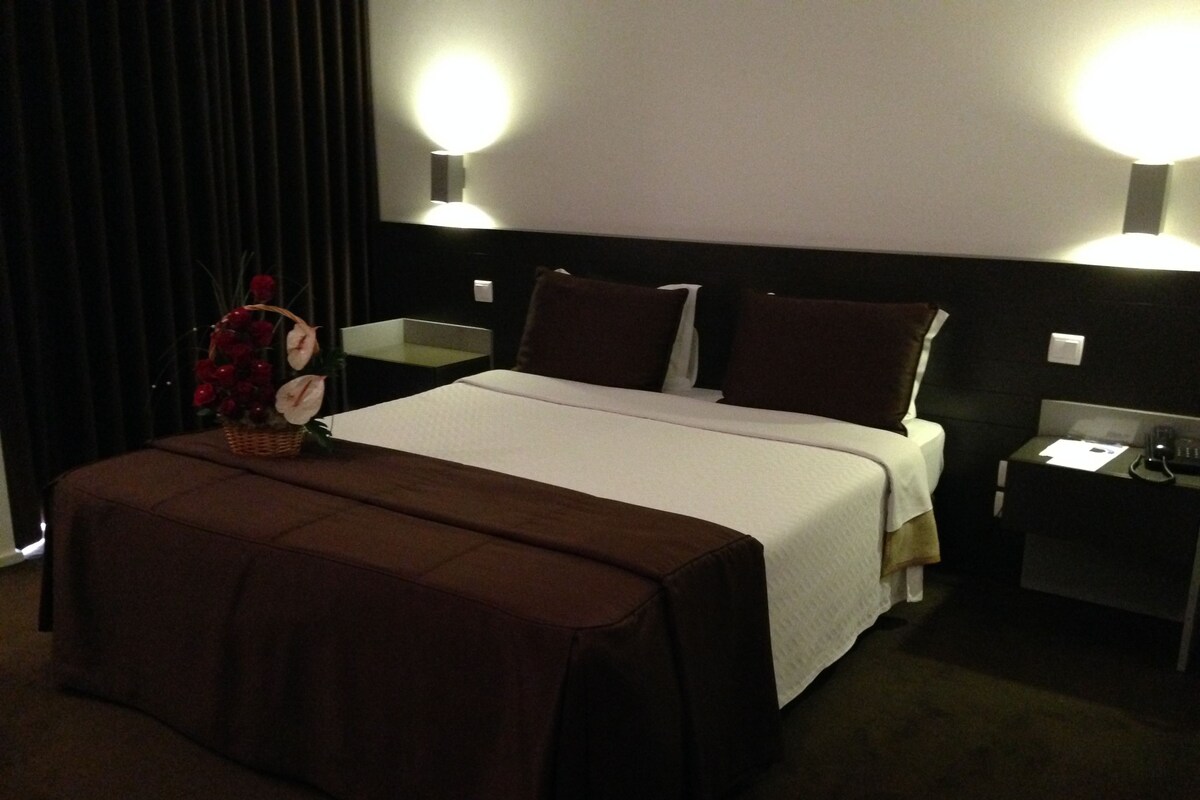 A hotel room with a large bed, two bedside tables, a nightstand, and a wall-mounted lamp.