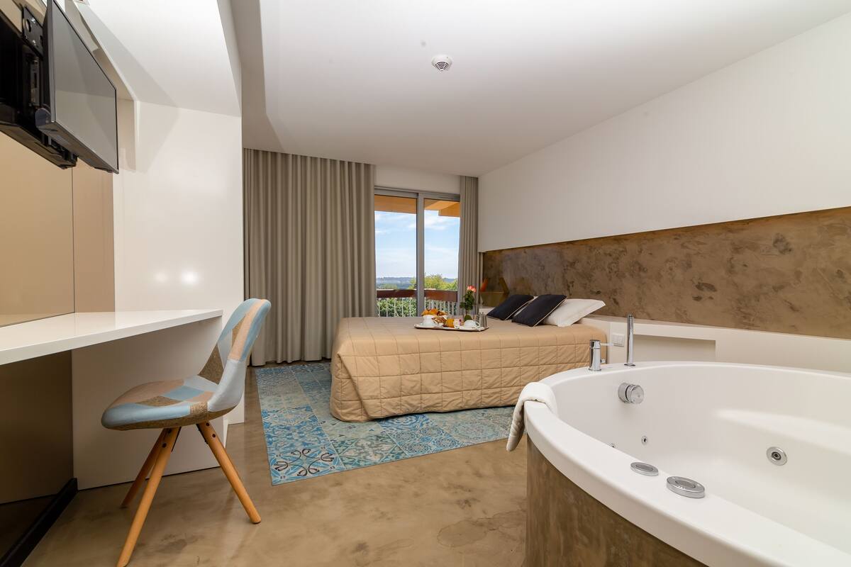 A modern hotel room with a bed, a desk, a chair, a large bathtub, and a view of the outdoors.