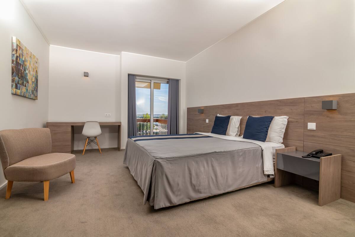 A modern hotel room with a large bed, a desk, a chair, and a view of the outdoors.