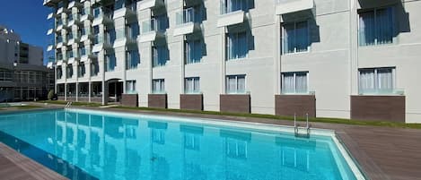 A modern hotel with a swimming pool, multiple balconies, and a clear blue sky.