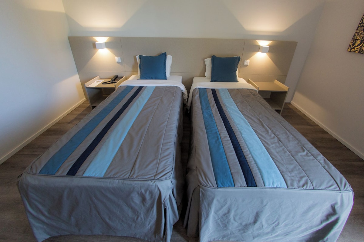 A hotel room with two single beds, each with a bedside table and lamp.