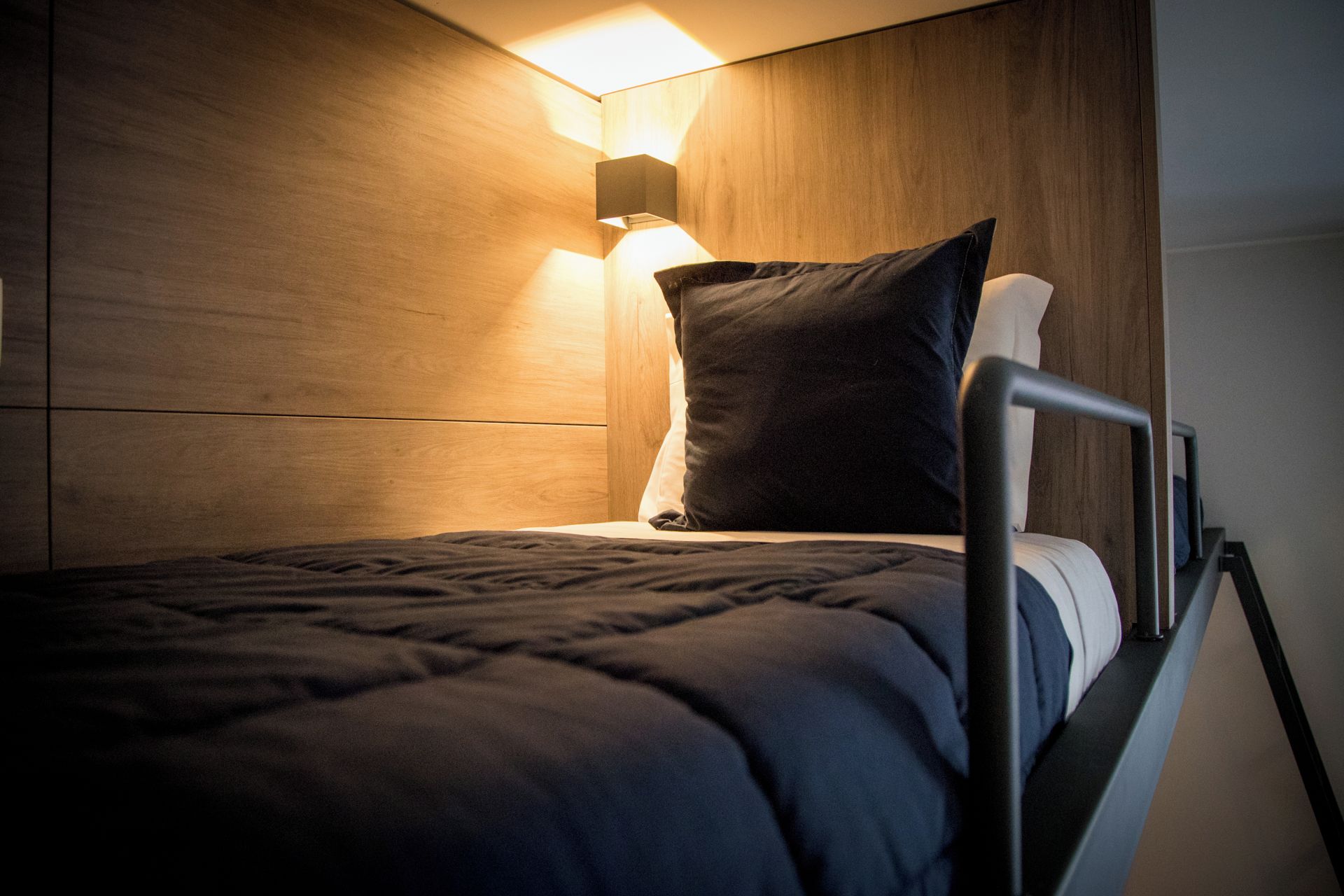 A single bed with a metal frame, a dark-colored mattress, and a pillow. A wall-mounted light fixture illuminates the bed area.
