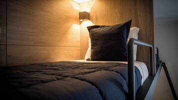A single bed with a metal frame, a dark-colored mattress, and a pillow. A wall-mounted light fixture illuminates the bed area.