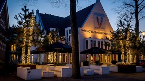 Un hotel con techo triangular y un letrero que dice "Parkhotel Brasserie Restaurant". Hay áreas de estar al aire libre con sombrillas y árboles decorados con luces.