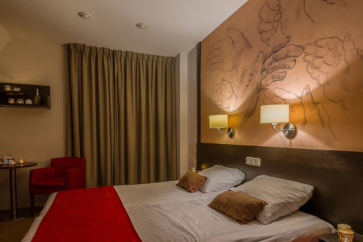 Une chambre d’hôtel avec un lit, une chaise rouge, un bureau et un mur muni d’un luminaire.