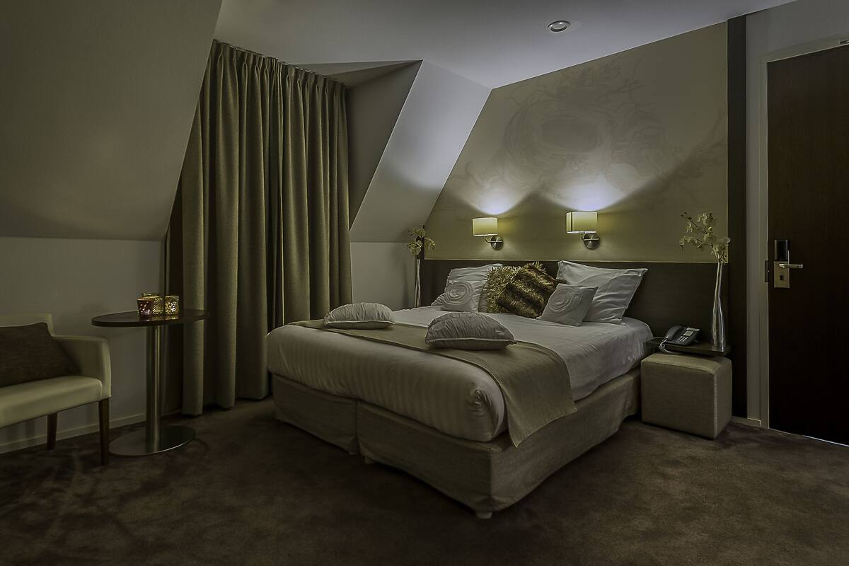 Une chambre d’hôtel moderne dotée d’un grand lit, d’une table de chevet avec une lampe, d’une petite table d’appoint et d’un coin salon avec un fauteuil.