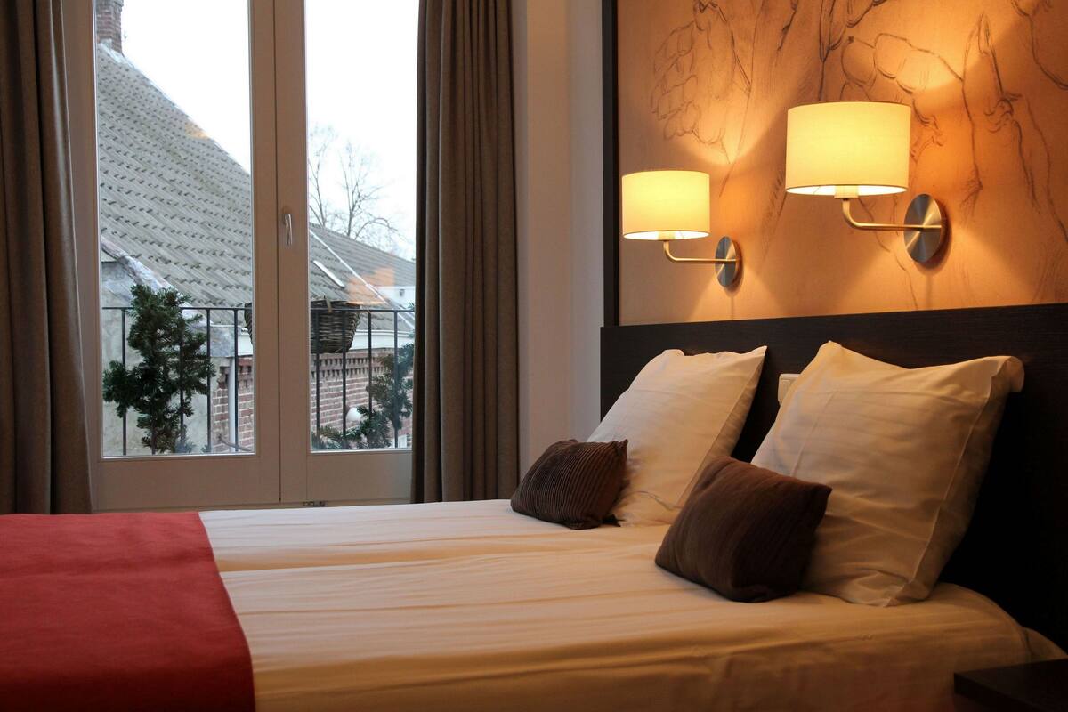 Une chambre d’hôtel avec un lit, deux lampes fixées au mur et une vue sur un immeuble par la fenêtre.
