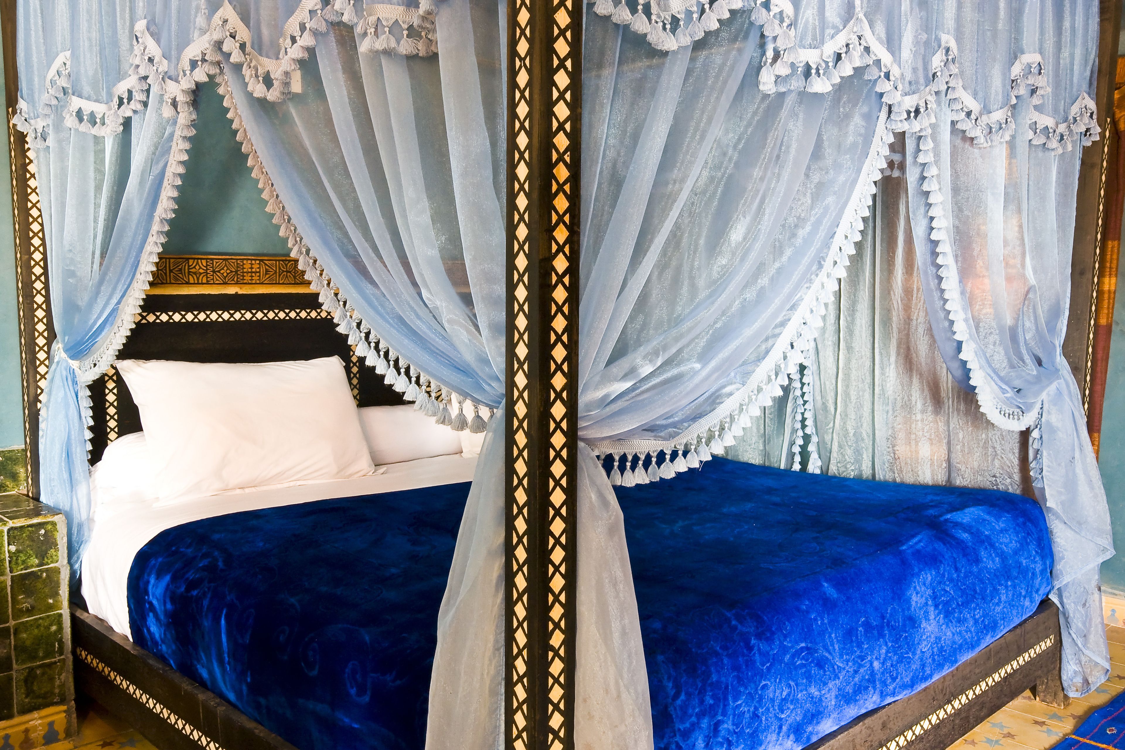 A four-poster canopy bed with a blue bedspread and white pillows, set against a blue wall with a decorative headboard.
