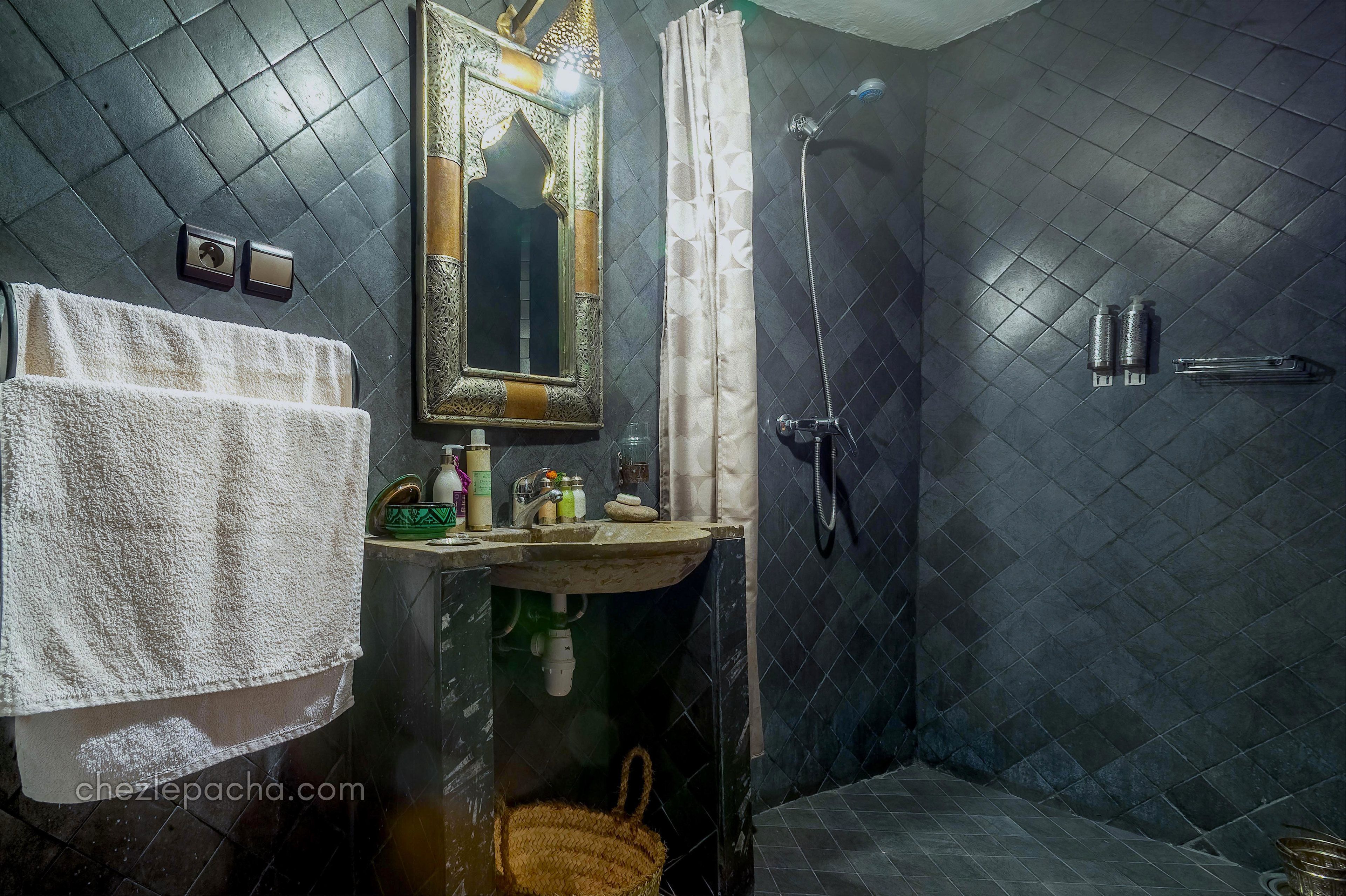 A bathroom with a dark tiled floor, a sink with toiletries, a mirror, and a shower area.