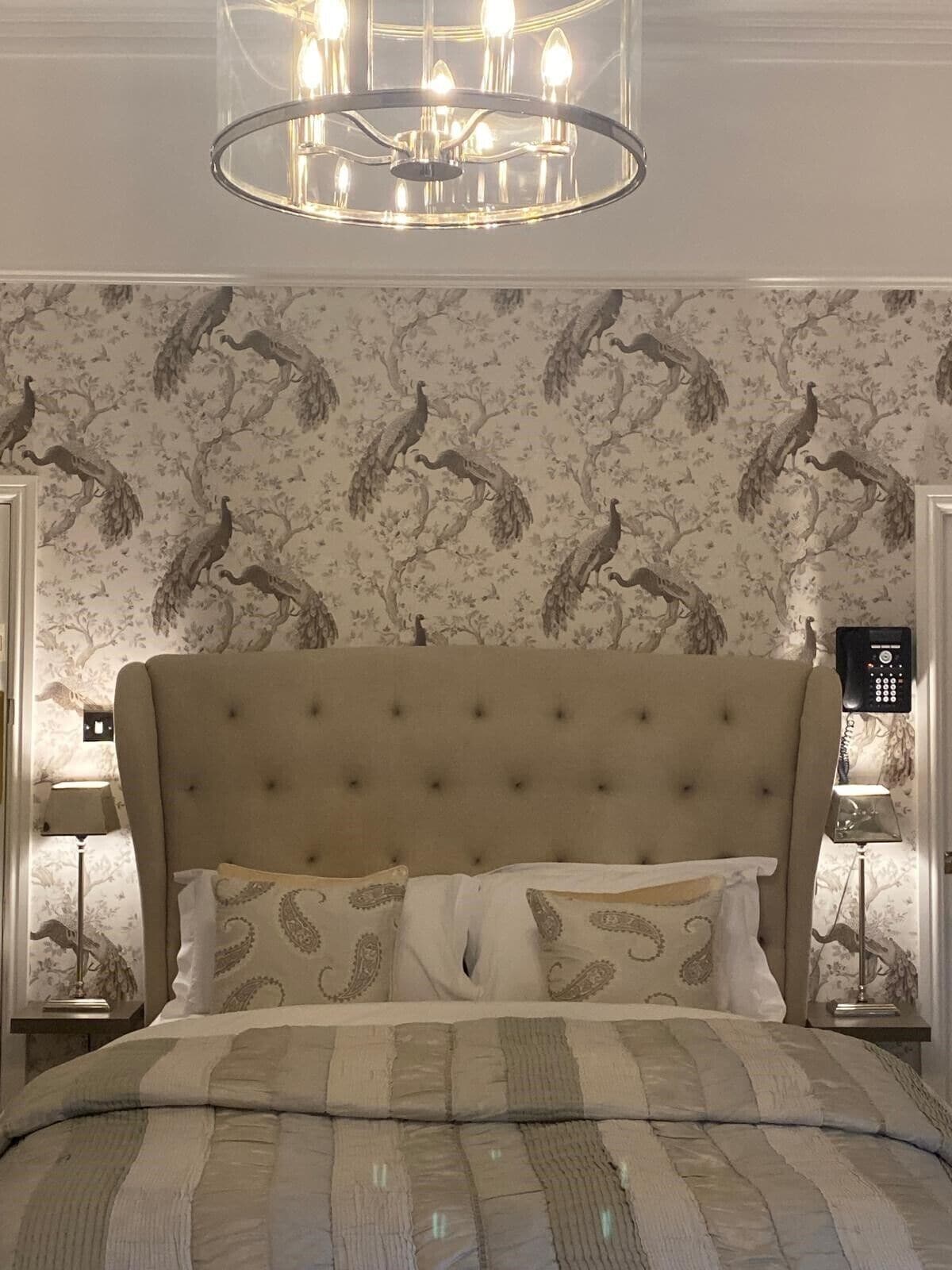 A bedroom with a tufted headboard, patterned wallpaper, a bed with multiple pillows, and a chandelier.