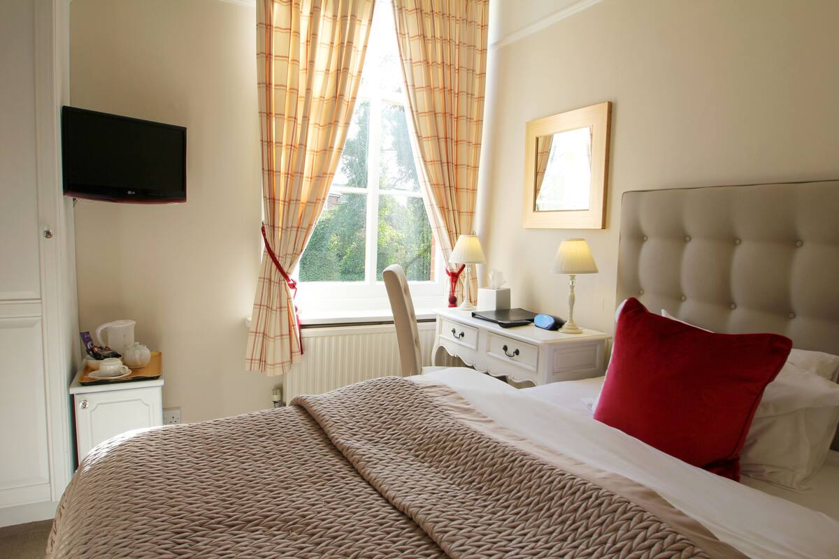 A hotel room with a bed, a television mounted on the wall, a window with curtains, a bedside table with a lamp, and a chair.