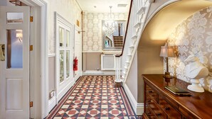 A hallway with patterned wallpaper, a geometric patterned floor, a wooden dresser with a lamp, and a staircase with a view to the upper floor. - The Alexandra Court Hotel (Congleton)