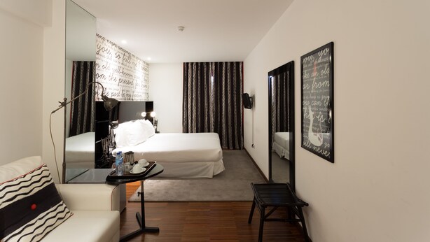 A hotel room with a bed, a desk with a chair, a mirror, and a wall with decorative text. - Porto Trindade Hotel (Porto)