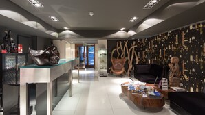 A modern interior with a reception desk, a leather chair, a coffee table with magazines, and a wall with abstract art. - Porto Trindade Hotel (Porto)