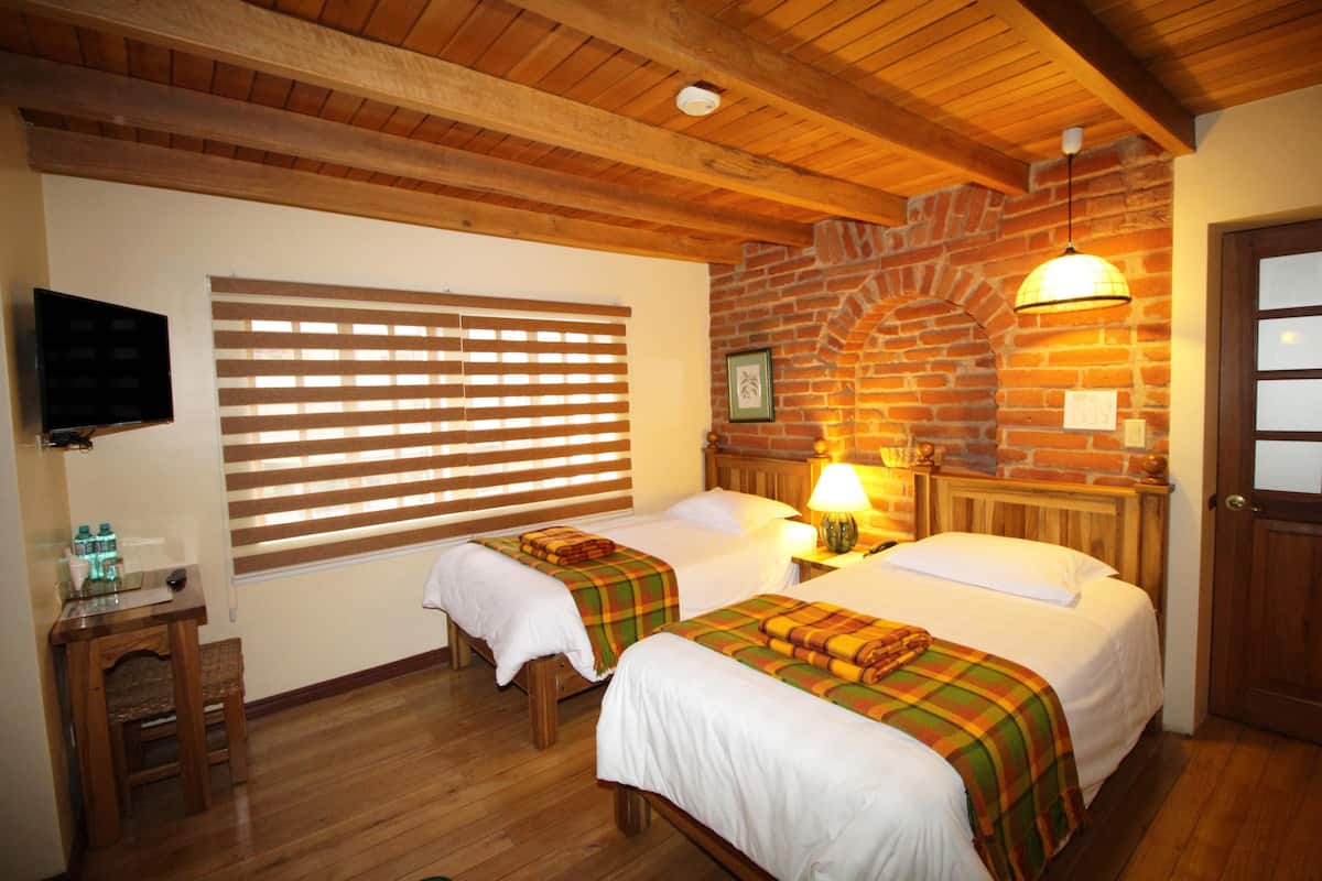 Una habitación con dos camas, suelo de madera, pared de ladrillo, televisión y una ventana con persianas.