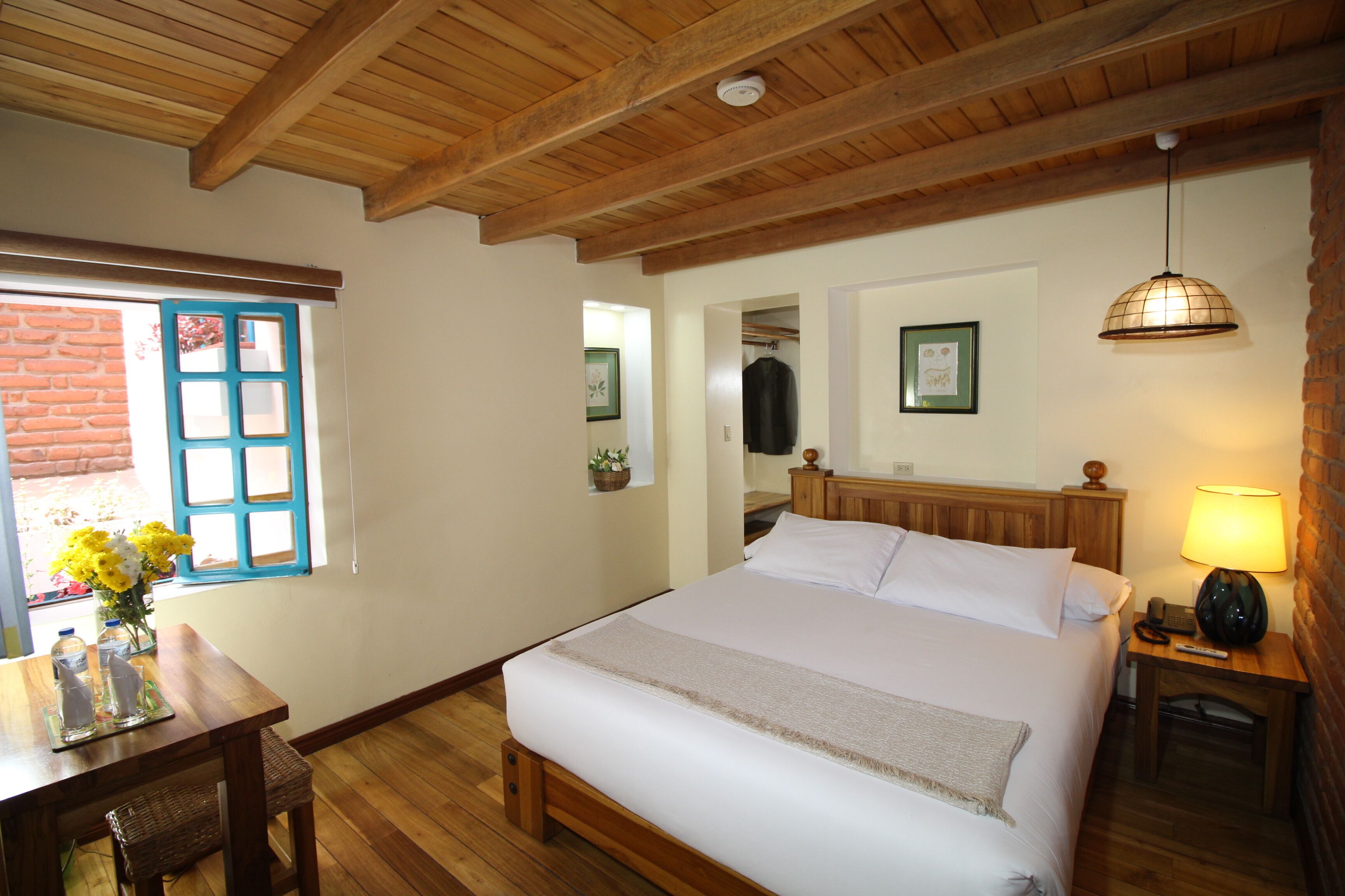 A bedroom with a wooden bed, a nightstand with a lamp, a window with blue shutters, and a wooden ceiling.