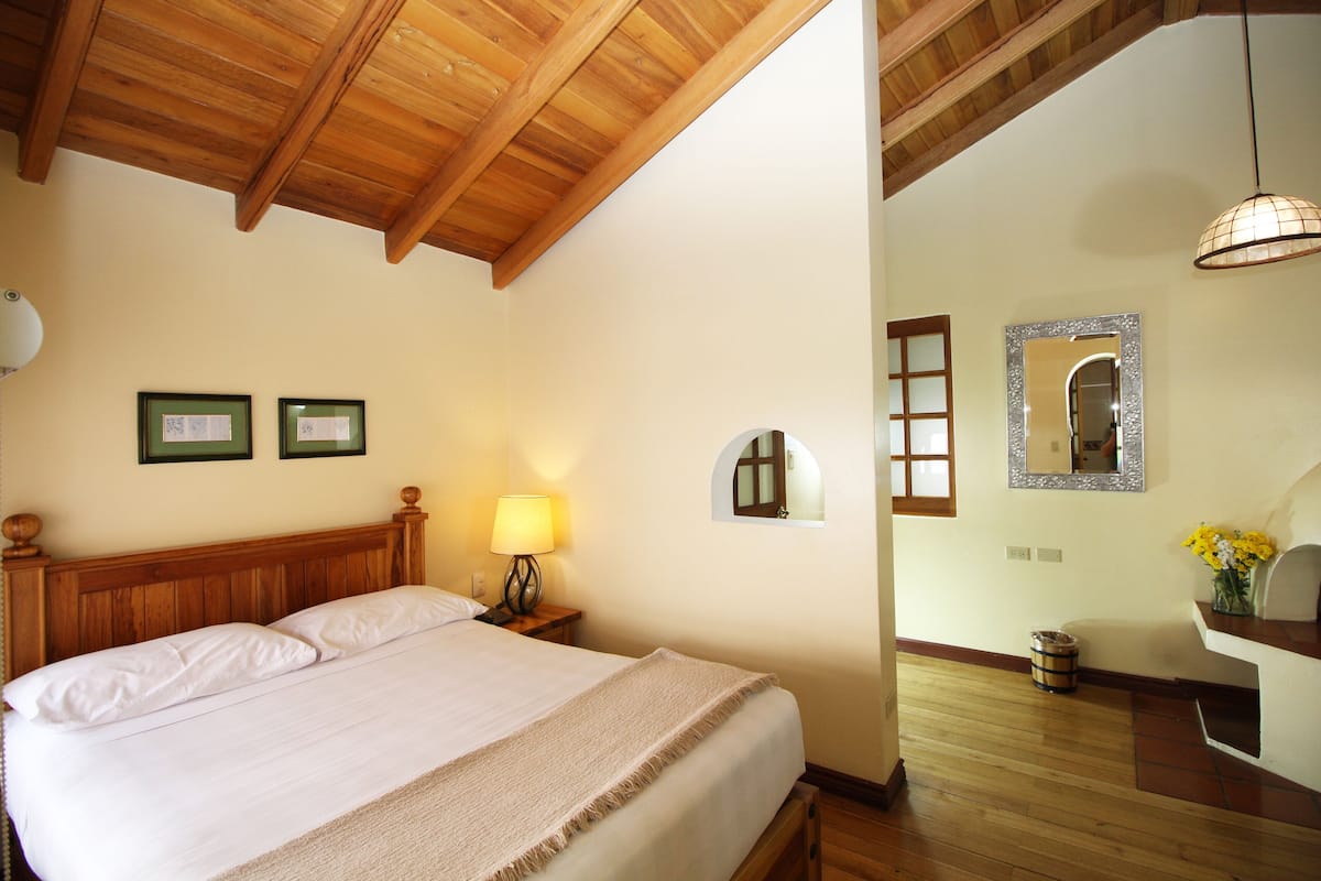 Un dormitorio con techo de madera, una cama con ropa blanca, una mesita de noche con lámpara, cuadros enmarcados en la pared y una ventana pequeña.