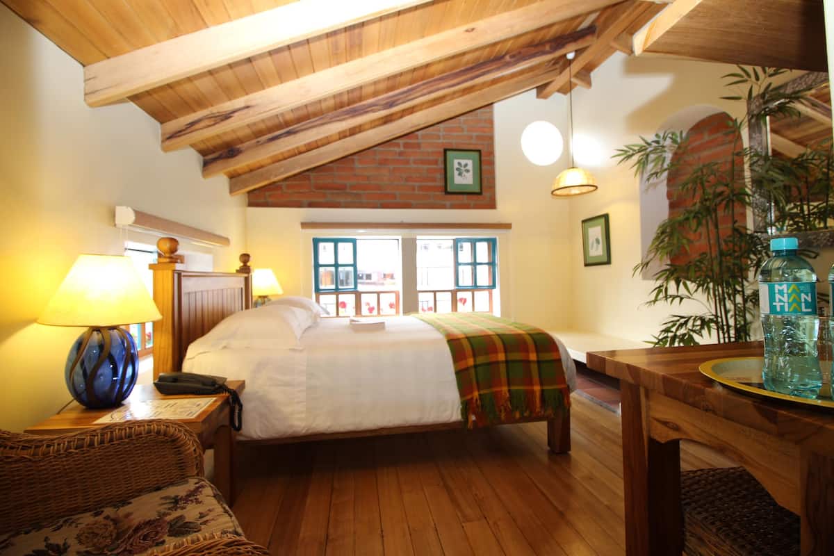 Un dormitorio con techo de madera, una cama con edredón blanco, una silla de mimbre, una mesita de noche con lámpara y una ventana con vistas.