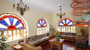 A cozy room with a wooden bench, a wicker chair, and a wooden table. There are two large windows with stained glass, a chandelier, and a birdcage. - Vieja Cuba Hotel (Quito)