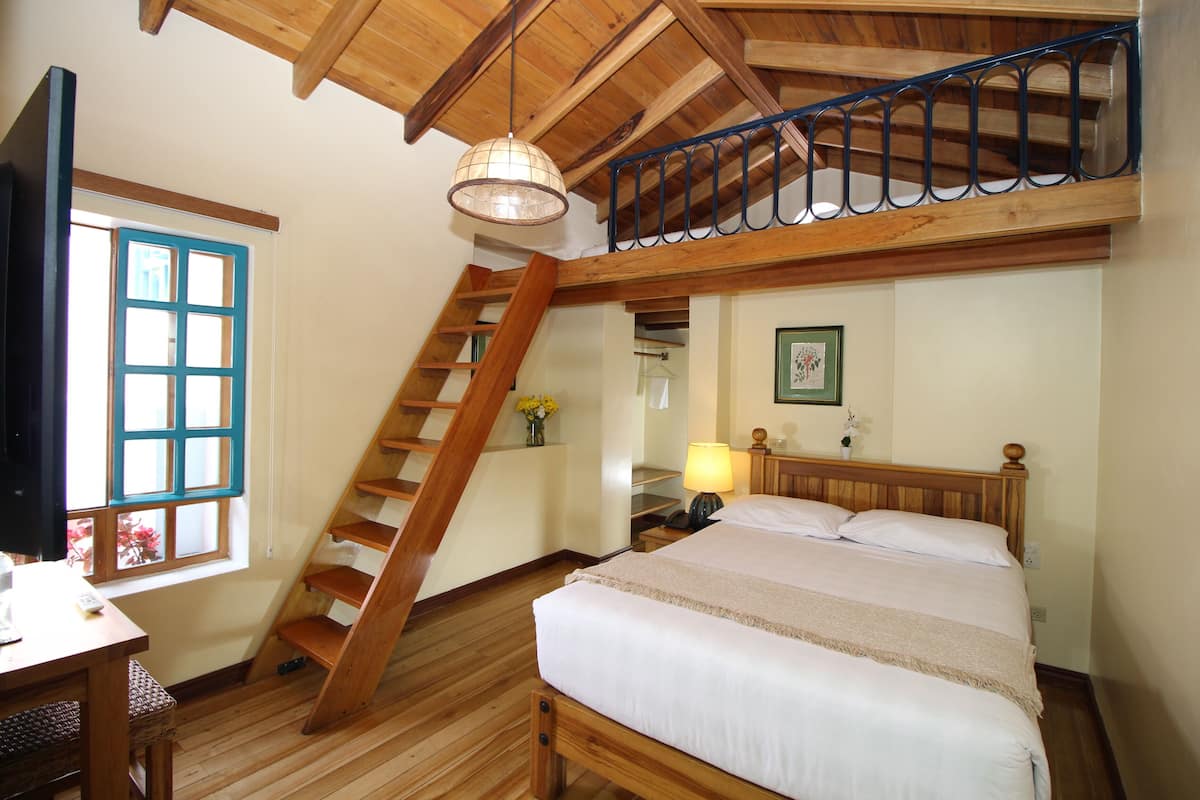 Un dormitorio con una cama de madera, un escritorio, una silla, un ventanal, una escalera y una lámpara colgante.