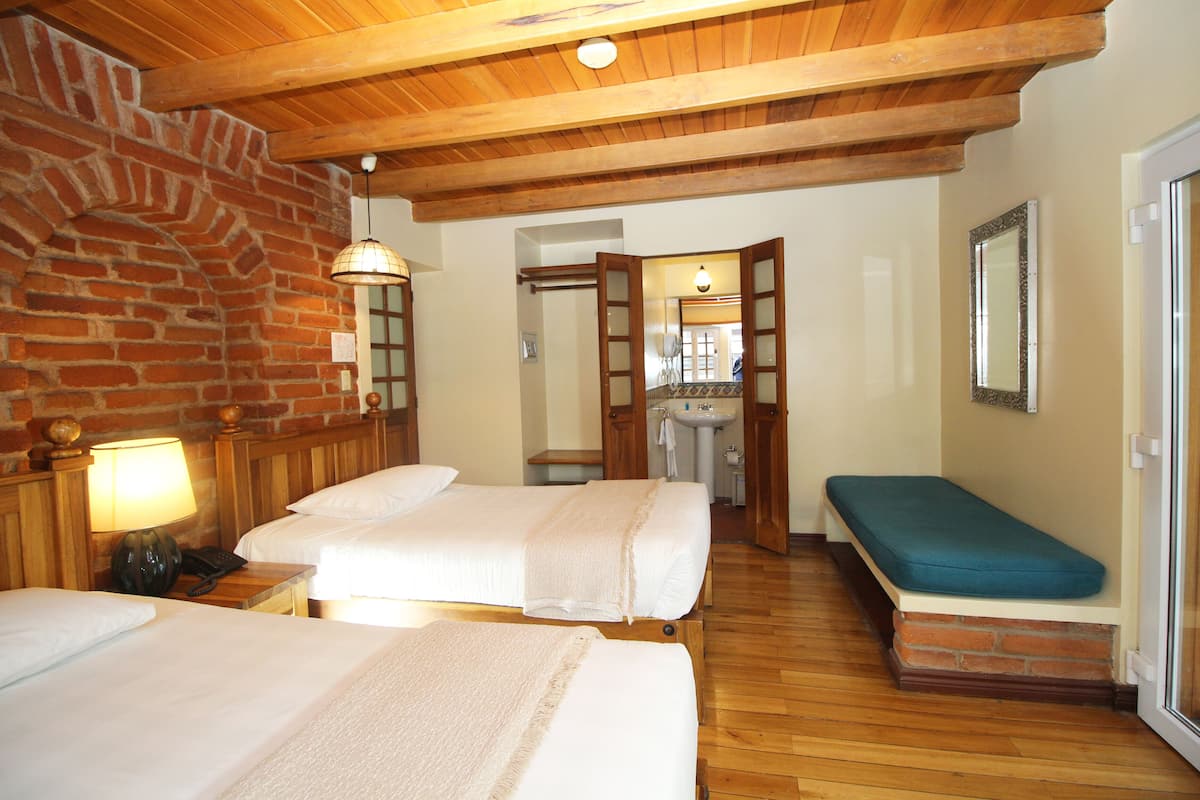 Una habitación con dos camas, un banco, pared de ladrillo, vigas de madera en el techo, un espejo y un lavabo.