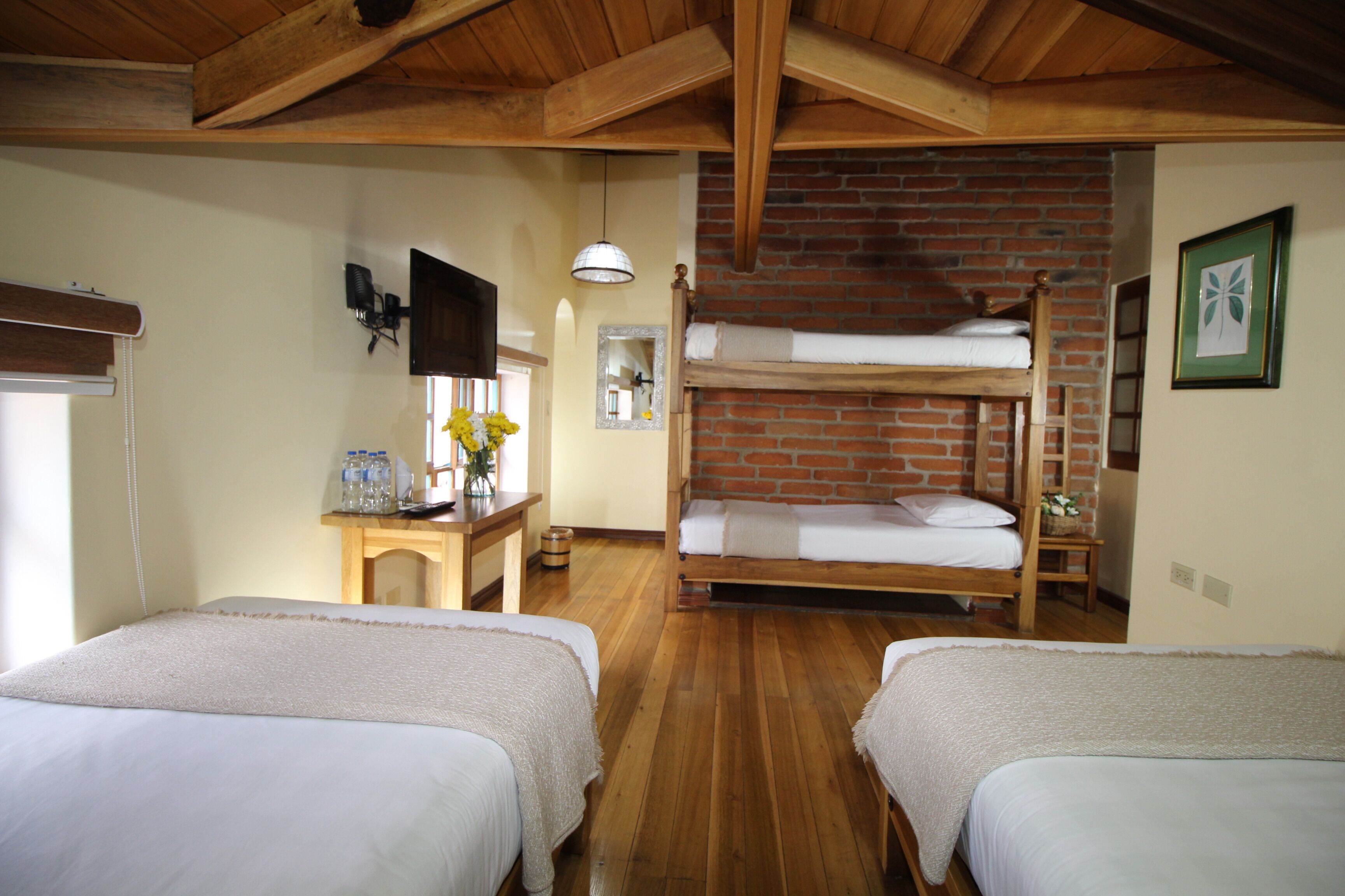 A room with two single beds, a bunk bed, wooden floors, a desk with a lamp, a mirror, and a framed picture on the wall.