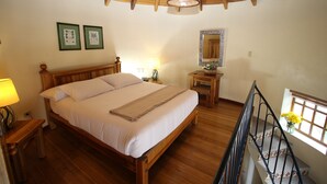 A bedroom with a wooden bed, a nightstand with a lamp, a small table with a vase of flowers, a mirror, and a window with a view of the outside. - Vieja Cuba Hotel (Quito)