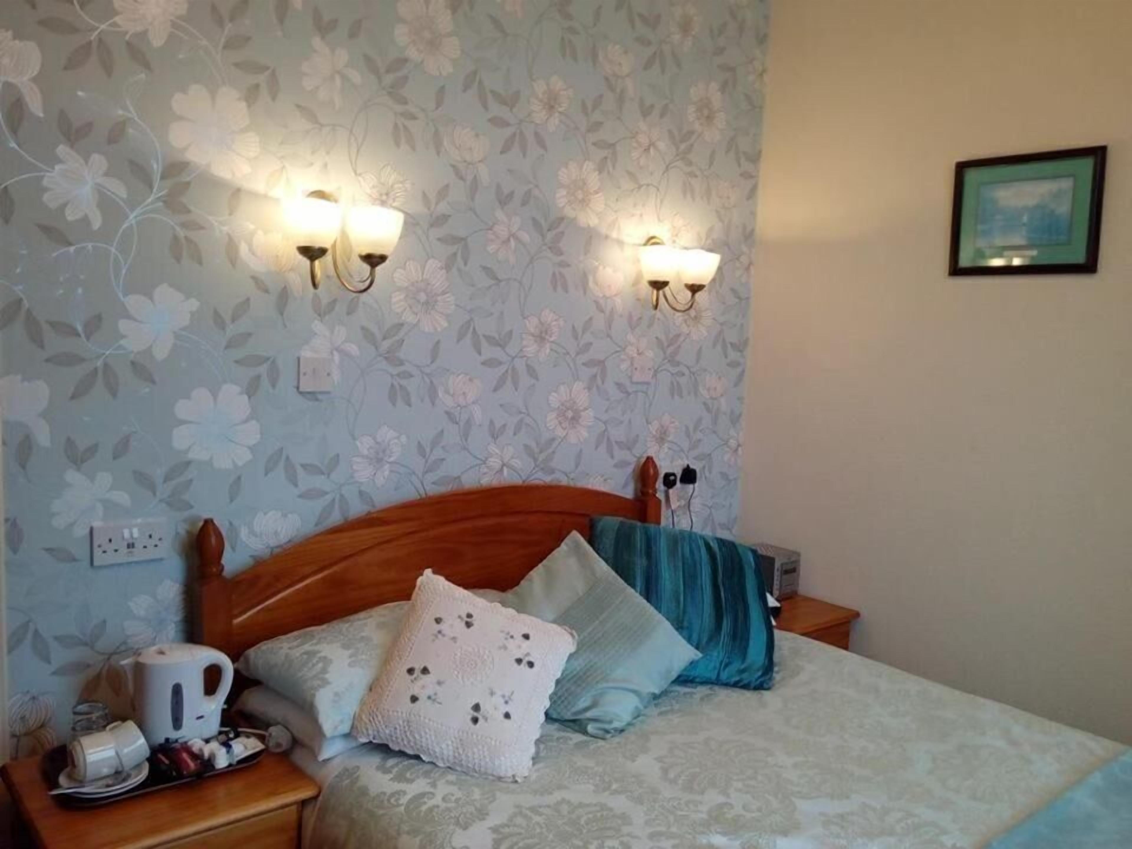 A bedroom with a floral wallpaper, a wooden headboard, two bedside lamps, a bed with patterned bedding, a bedside table with a kettle and cups, and a framed picture on the wall.