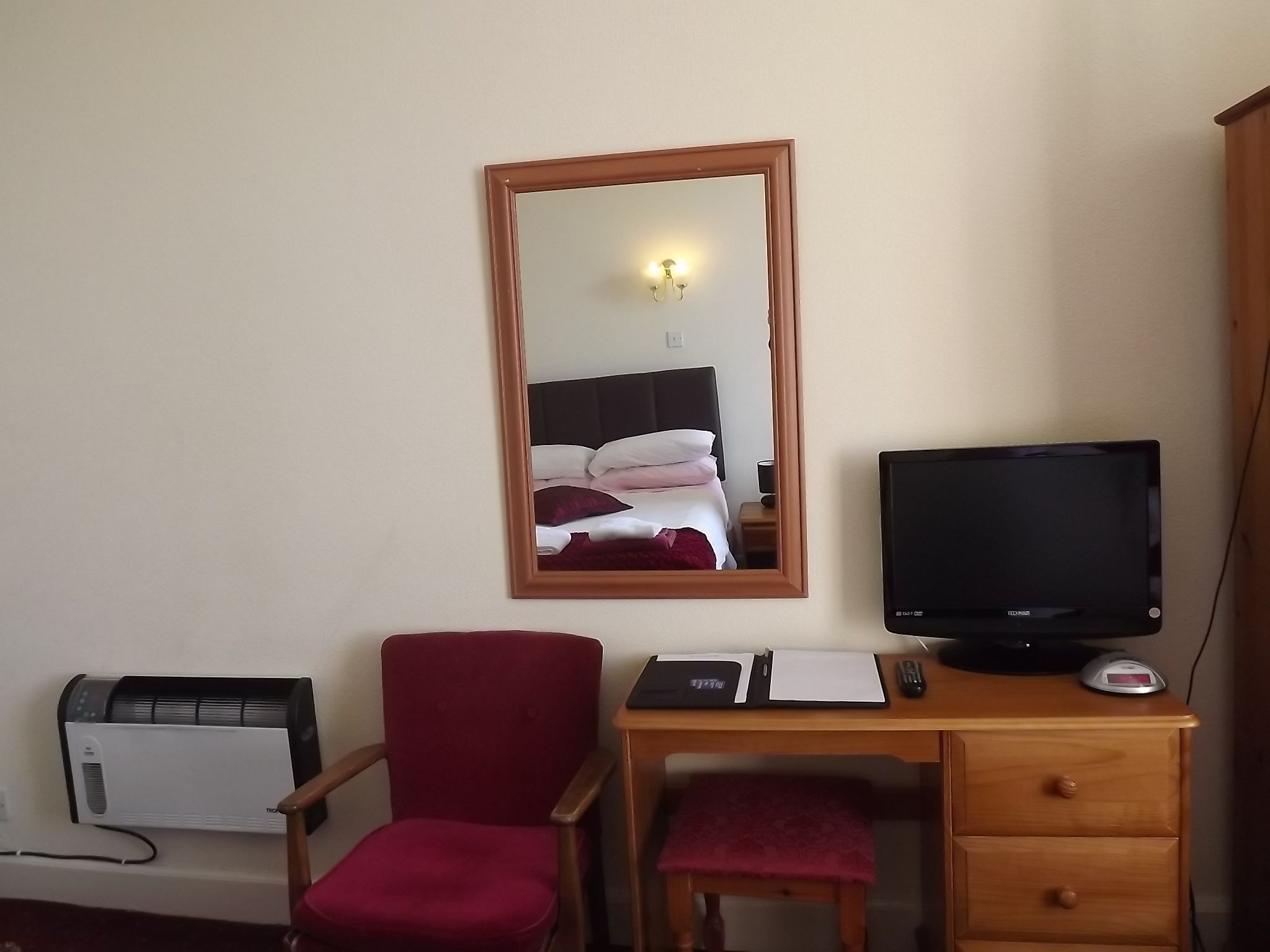 A hotel room with a wooden desk, a red chair, a television, a mirror, and a heater.