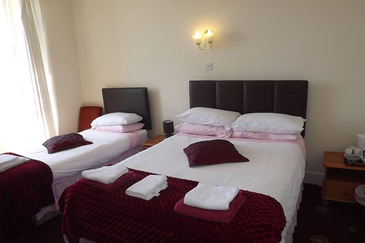 Quarto de hotel com duas camas, cada uma com lençóis brancos e cobertores vermelhos. Há luminárias fixadas na parede acima das camas e uma pequena mesa com um abajur e uma xícara.